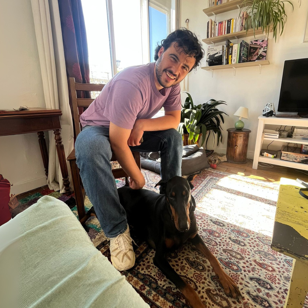 Un homme souriant, accroupi à côté d’un Dobermann noir et feu, dans un salon lumineux avec un tapis coloré et une bibliothèque murale.