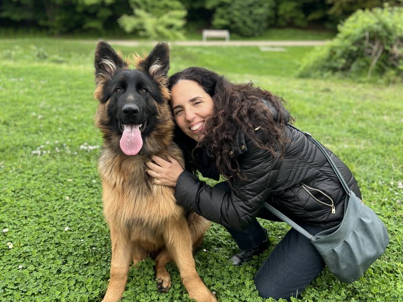 Berger allemand adulte assis sur de l’herbe aux côtés d’une femme aux cheveux bouclés, souriante, en extérieur.