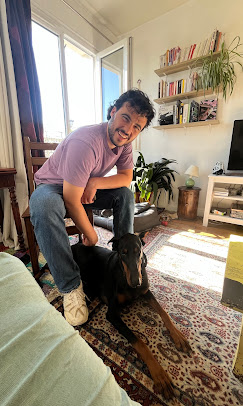 Un homme souriant, accroupi à côté d’un Dobermann noir et feu, dans un salon lumineux avec un tapis coloré et une bibliothèque murale.